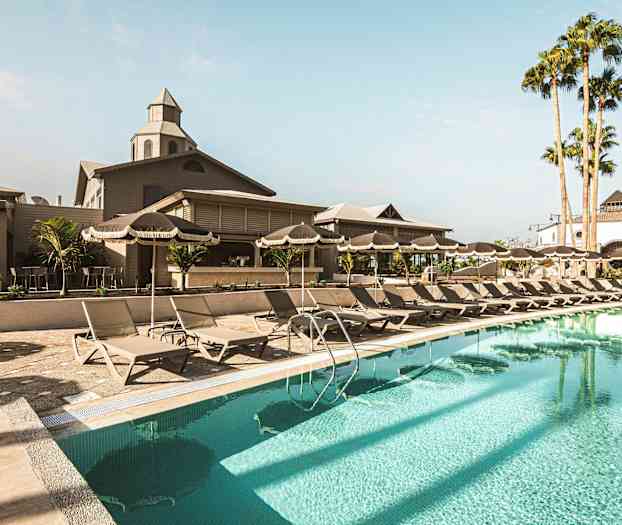 Billede af et poolområde med liggestole, parasoller og en bygning i baggrunden. (AI-genereret tekst)