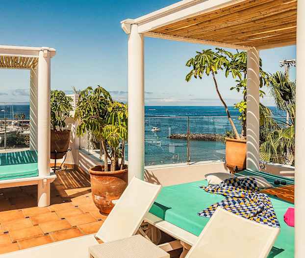 En solrig terrasse med udsigt over havet. En dagseng med turkis madras og farverige stoffer står under en pergola. Rundt terrassen er der potteplanter og palmer. (AI-genereret tekst)