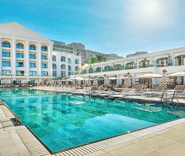 En hotelbygning med en stor swimmingpool og liggestole foran. (AI-genereret tekst)