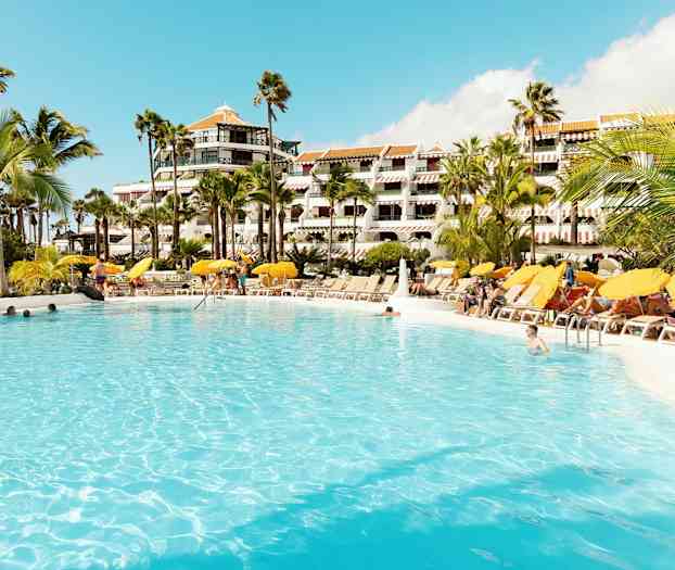 Stor swimmingpool omgivet af liggestole og parasoller foran hotelbygning. (AI-genereret tekst)