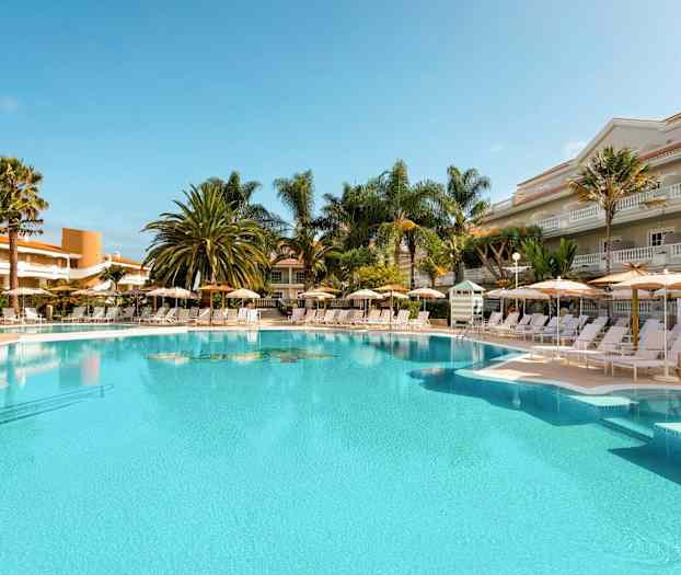 Udendørspool med liggestole og parasoller omgivet af palmer og bygninger. (AI-genereret tekst)