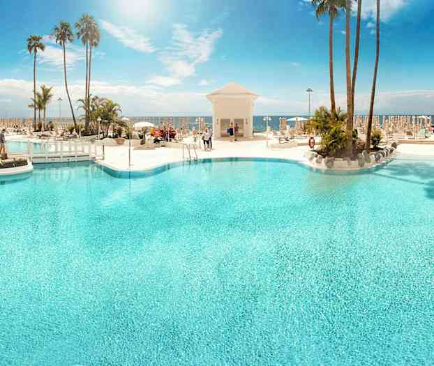 En stor udendørs pool omgivet af liggestole og palmer med havet i baggrunden. (AI-genereret tekst)