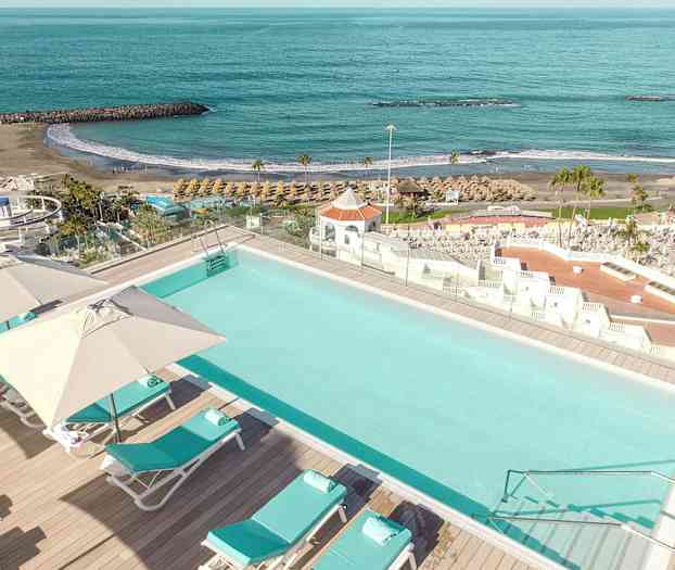 En pool med liggestole og parasoller, beliggende tæt på en strand og havet. (AI-genereret tekst)