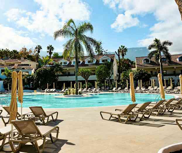 Et hotelkompleks med en swimmingpool, liggestole og parasoller. Bygninger omgivet af palmetræer. (AI-genereret tekst)