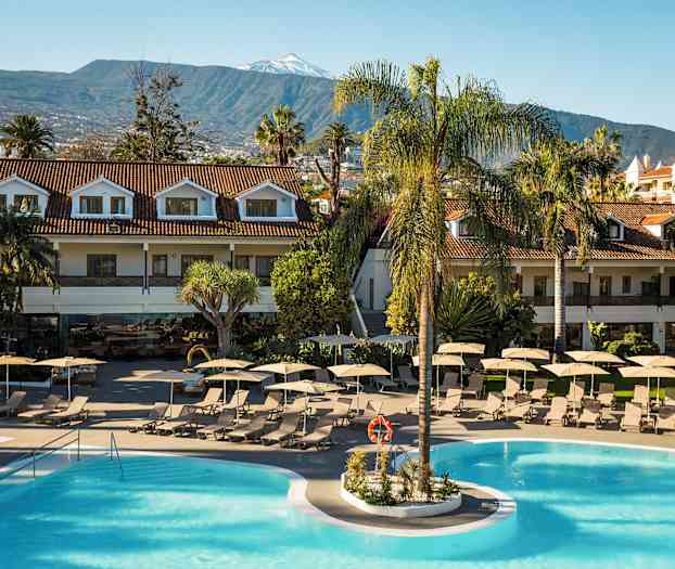 Et resort med poolområde omgivet af liggestole og parasoller. Tilhørende bygninger med røde tage og bjerge i baggrunden. (AI-genereret tekst)