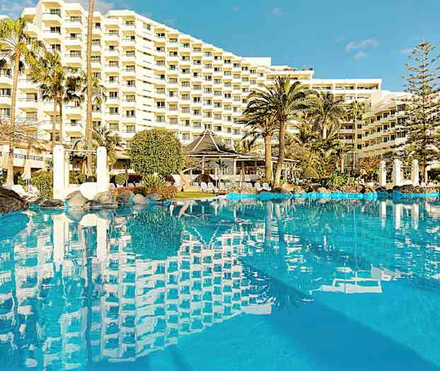 Et billede af en udendørs pool omgivet af palmer og et stort hotel i baggrunden. (AI-genereret tekst)