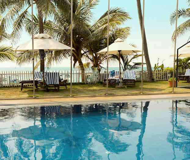 En udendørs pool med palmer og solsenge i baggrunden samt havudsigt. (AI-genereret tekst)