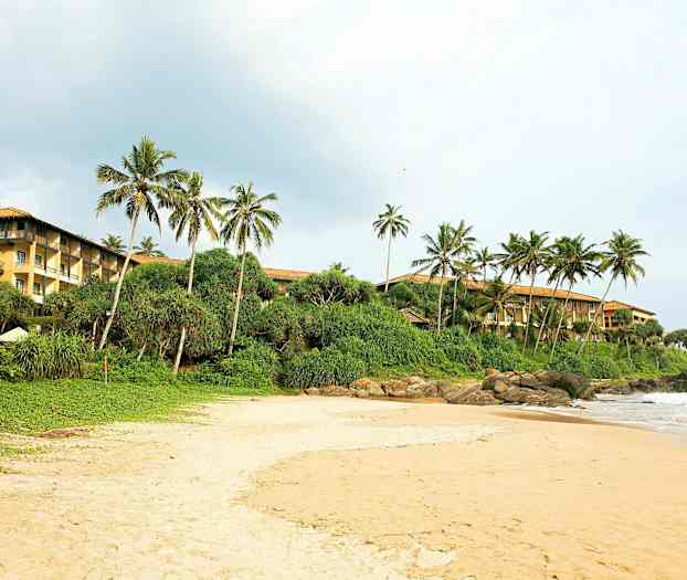 En sandstrand med grøn buskads og palmer i baggrunden. Til højre ses en bygning med gule vægge. (AI-genereret tekst)