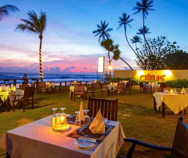 En udendørs restaurant med hvide duge og træstole ved solnedgang nær havet. (AI-genereret tekst)