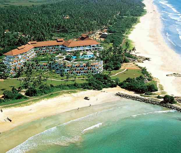 Luftfoto af et hotelkompleks omgivet af grøntområder ved kysten med en lang sandstrand og hav. (AI-genereret tekst)