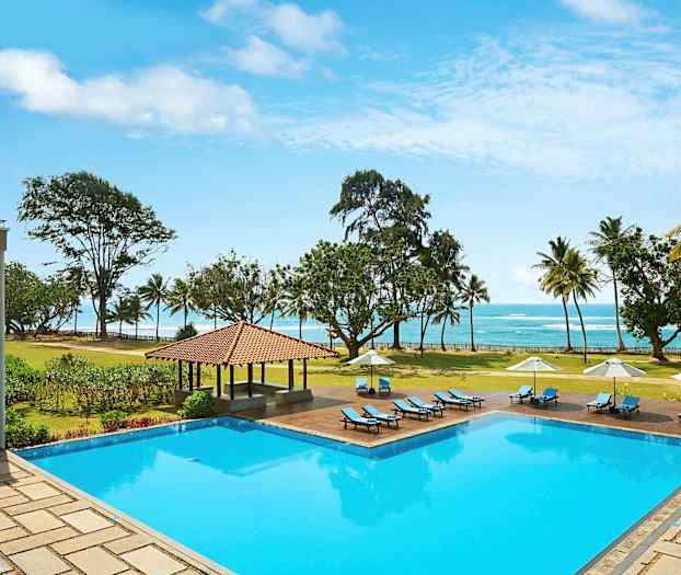 En udendørs swimmingpool med liggestole og parasoller, omgivet af en græsplæne og træer, med udsigt over stranden og havet. (AI-genereret tekst)