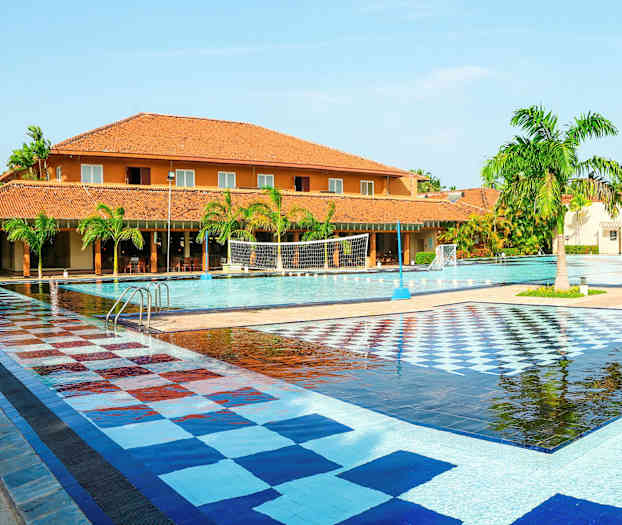 En stor udendørs pool med flisebelagt farverigt område, omgivet af palmer og bygninger med teglfarvede tage. (AI-genereret tekst)