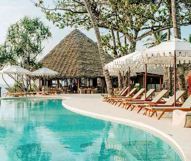 Et poolområde med liggestole og parasoller. I baggrunden ses en bygning med stråtag og palmer. (AI-genereret tekst)