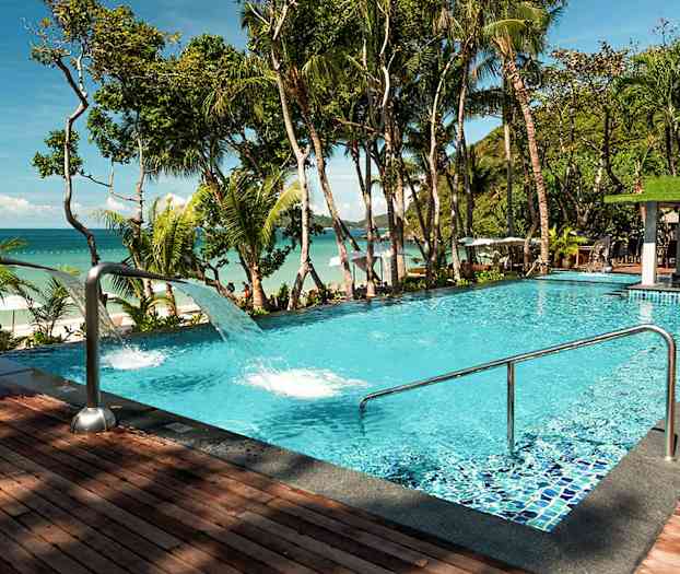 En pool med klart blåt vand, omgivet af tropiske træer og med udsigt over havet. (AI-genereret tekst)