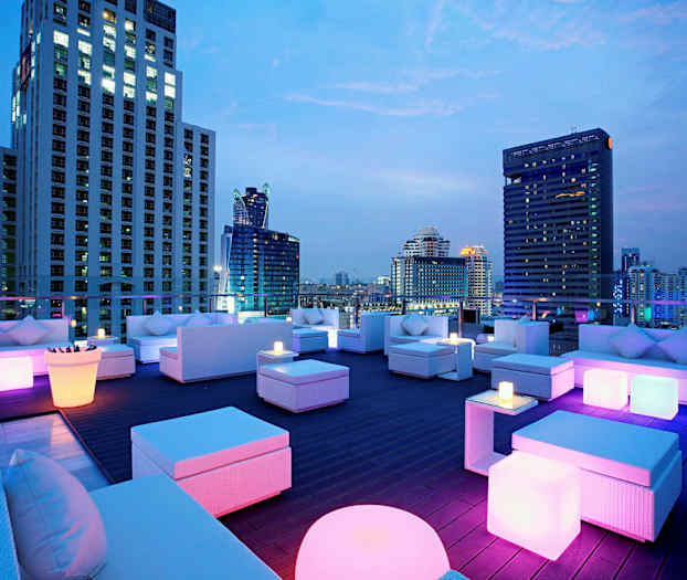 En tagterrasse med moderne møbler og farverig belysning, omgivet af høje bygninger om aftenen. (AI-genereret tekst)