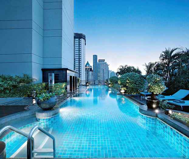 En udendørs pool ved en bygning omkranset af beplantning, med liggestole og høje bygninger i baggrunden. (AI-genereret tekst)