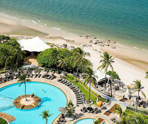 Billede af en pool med liggestole omgivet af grønne områder, beliggende ved siden af en sandstrand, der møder havet. (AI-genereret tekst)