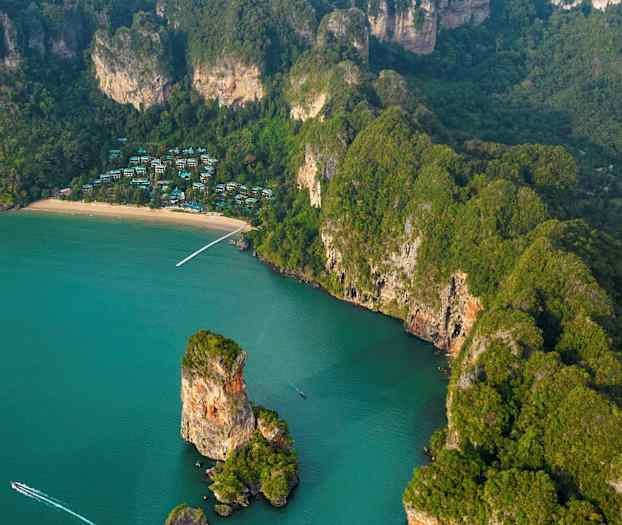 Luftfoto af en tropisk kyst med en strand, klipper og grøn vegetation, samt huse synlige mellem træerne. (AI-genereret tekst)