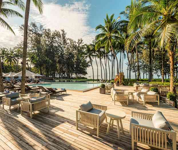 En träterrasse med grå sofaarrangementer og sofaborde, liggestole omkring en pool samt omgivelser af palmer og træer. (AI-genereret tekst)