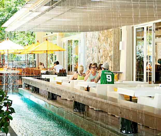 Et udendørs område med borde og stole, samt en vandkanal og gulhvide parasoller. (AI-genereret tekst)