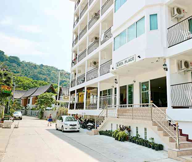 Udenfor Kata Place, en moderne bygning med altaner, står parkerede køretøjer, og en kvinde passerer forbi. (AI-genereret tekst)