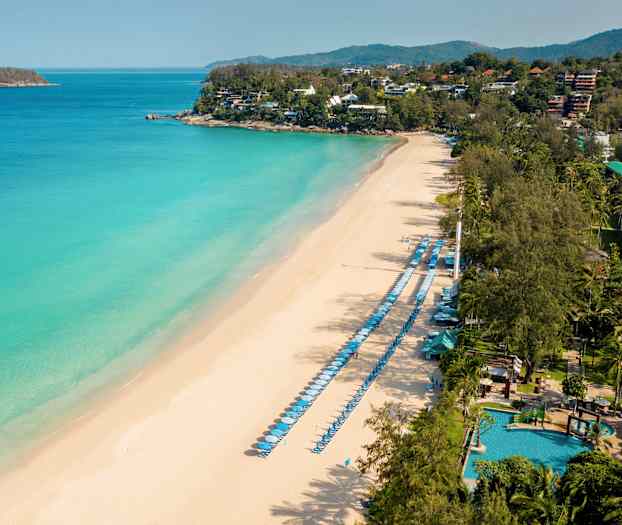 Luftfoto af en sandstrand med liggestole og parasoller langs havet samt grønne områder i baggrunden. (AI-genereret tekst)