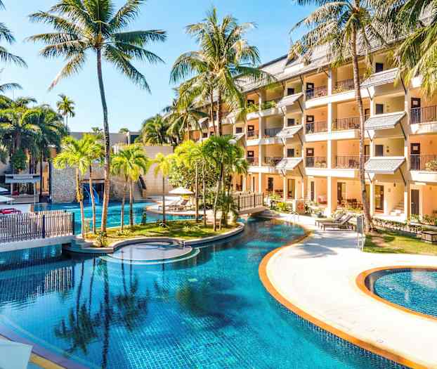 Billede af et hotelområde med en stor pool omgivet af palmer og bygninger. (AI-genereret tekst)