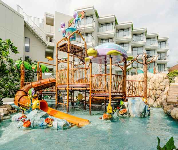 Legeplads med vandrutsjebane og legeredskaber, omgivet af grønne planter og stenblokke. Hotelbygning i baggrunden. (AI-genereret tekst)