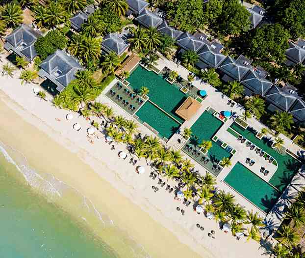 Luftfoto af en strand med parasoller, palmer, poolområde og bygninger. (AI-genereret tekst)