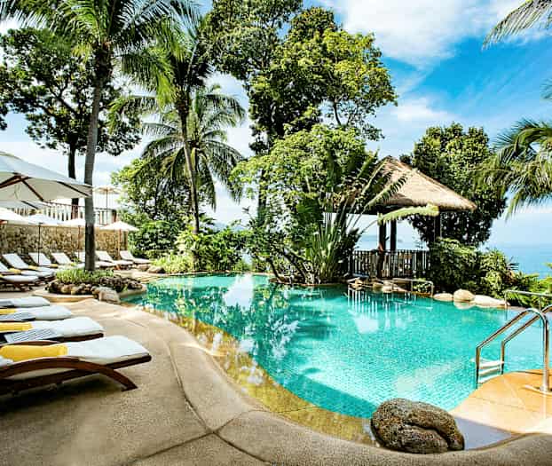 En udendørs swimmingpool omgivet af palmer, liggestole og parasoller, med udsigt over havet i baggrunden. (AI-genereret tekst)