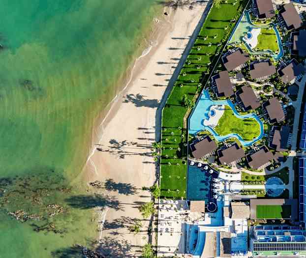 Luftfoto af en strand og et hotelområde med pools og bygninger. (AI-genereret tekst)