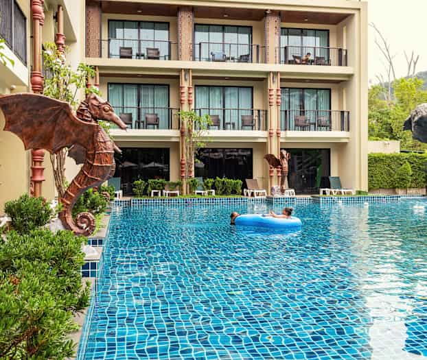 Udendørspool med et barn, der flyder på en blå badering, omgivet af hotelbygninger og grøntområder. (AI-genereret tekst)