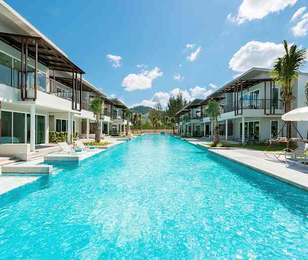 Udsigt over en lang pool omgivet af moderne villaer og liggestole under parasoller. (AI-genereret tekst)