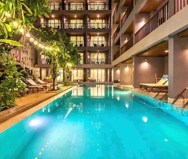 En udendørs pool omgivet af liggestole og grønne planter foran et moderne hotel. (AI-genereret tekst)