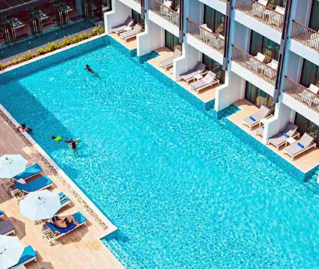 En udendørs pool med liggestole og parasoller ved siden af samt bygninger med terrasser. (AI-genereret tekst)