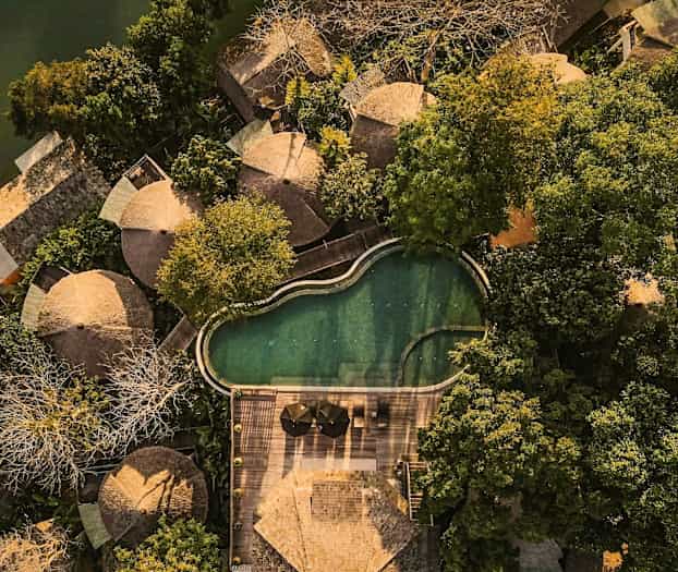 Luftfoto af flere runde huse omgivet af frodig vegetation, ved siden af en uregelmæssig pool og en træterrasse. (AI-genereret tekst)