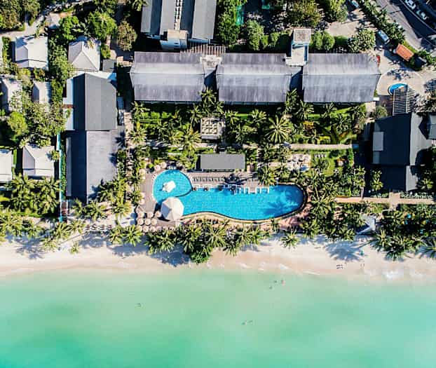 Luftfoto af en ferieresort med pool, omgivet af tropiske træer og tæt på en strand med klart vand. (AI-genereret tekst)