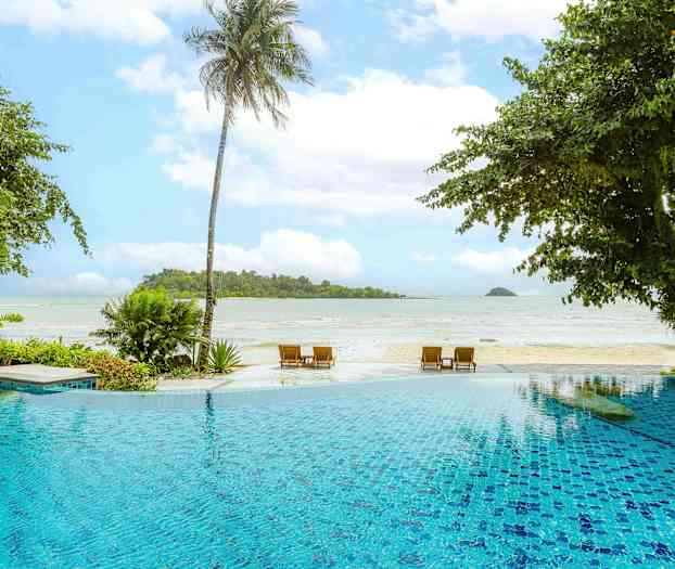 Et view over en udendørs pool i forgrunden med en strand og havet i baggrunden, omgivet af frodige planter og palmer. (AI-genereret tekst)
