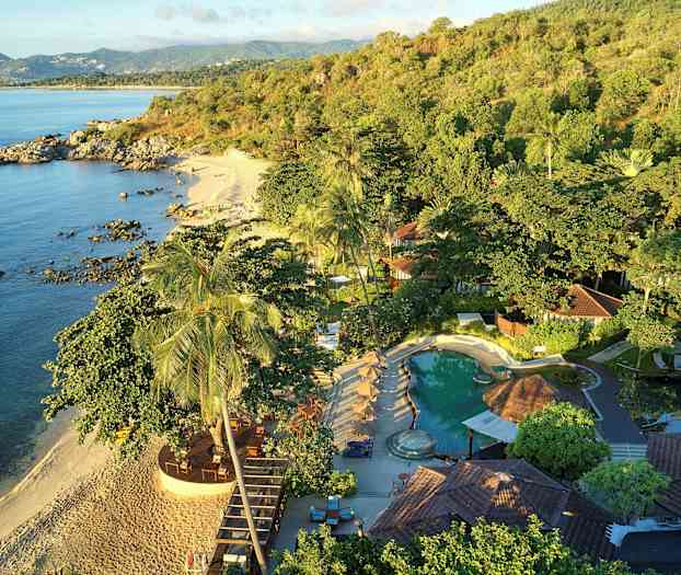 Billede, der viser en tropisk strand, omgivet af frodig vegetation og med en pool og bygninger i forgrunden. (AI-genereret tekst)