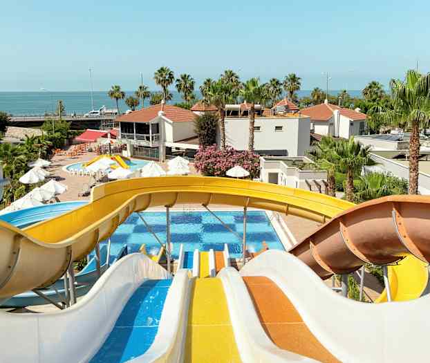 Flere farverige vandrutsjebaner fører ned til en pool med palmer og bygninger i baggrunden. (AI-genereret tekst)