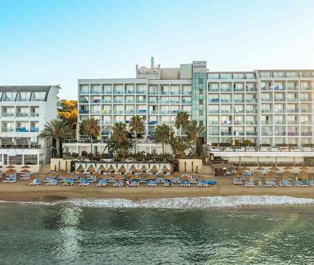 Billede af en stor hvid hotelbygning ved en solrig strand med liggestole og parasoller. (AI-genereret tekst)