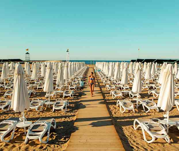 En kvinde i bikini går på en træsti mellem liggestole og parasoller på en strand. (AI-genereret tekst)