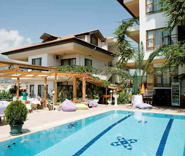 En bygning med hvid facade, omgivet af grønne planter og med en pool i forgrunden. (AI-genereret tekst)