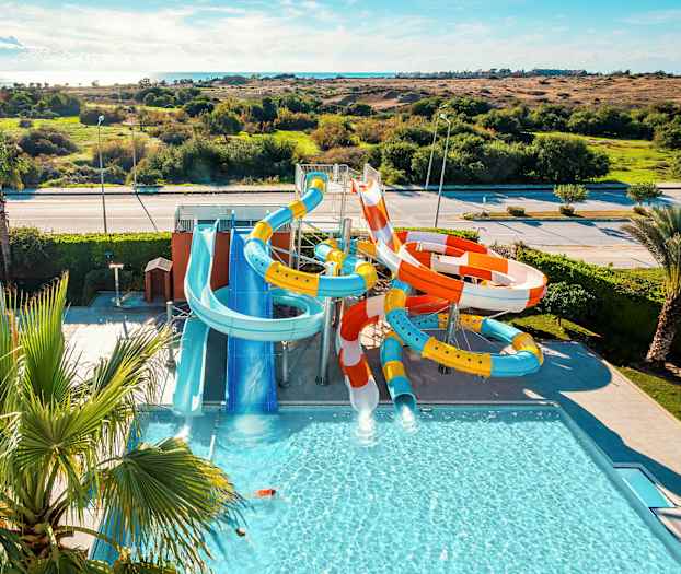 Billedet viser en vandpark med flere farvestrålende vandrutsjebaner og en pool, omgivet af græs og landskab. (AI-genereret tekst)