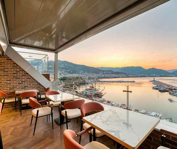 Restaurant med udsigt over en havn, havet og bjergene ved solnedgang. (AI-genereret tekst)