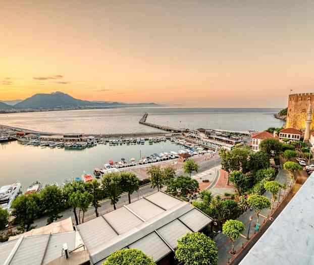 Billedet viser en havn med både og bygninger ved havet under solnedgangen. En borg ses på højre side. (AI-genereret tekst)