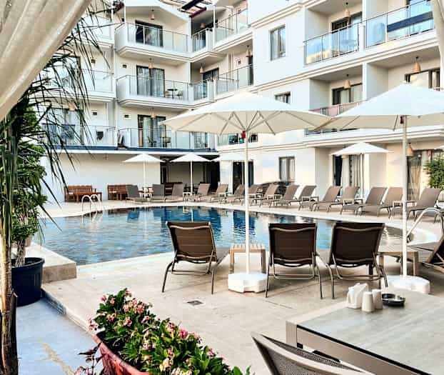 Billedet viser en swimmingpool ved et hotel omgivet af liggestole, parasoller og altaner i baggrunden. (AI-genereret tekst)