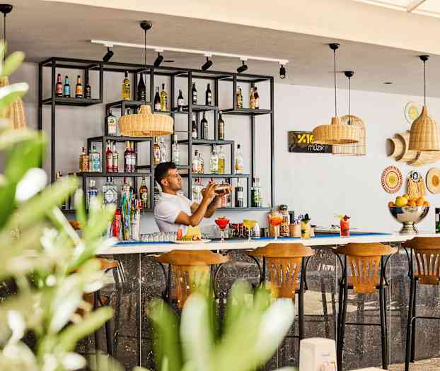 En bar med hylder fyldt med flasker og tre træstole foran. (AI-genereret tekst)