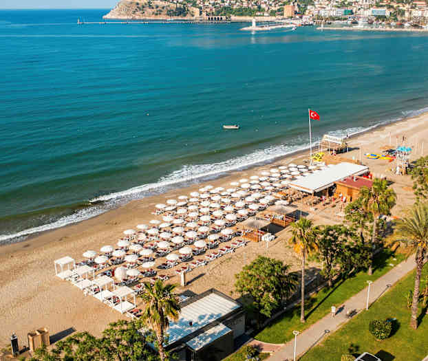 Luftfoto af en strand med liggestole og parasoller, omgivet af turkis hav og bygninger i baggrunden. (AI-genereret tekst)