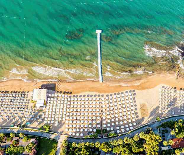 Et luftfoto af en strand med parasoller og solsenge samt en bro, der strækker sig ud i turkis vand. (AI-genereret tekst)